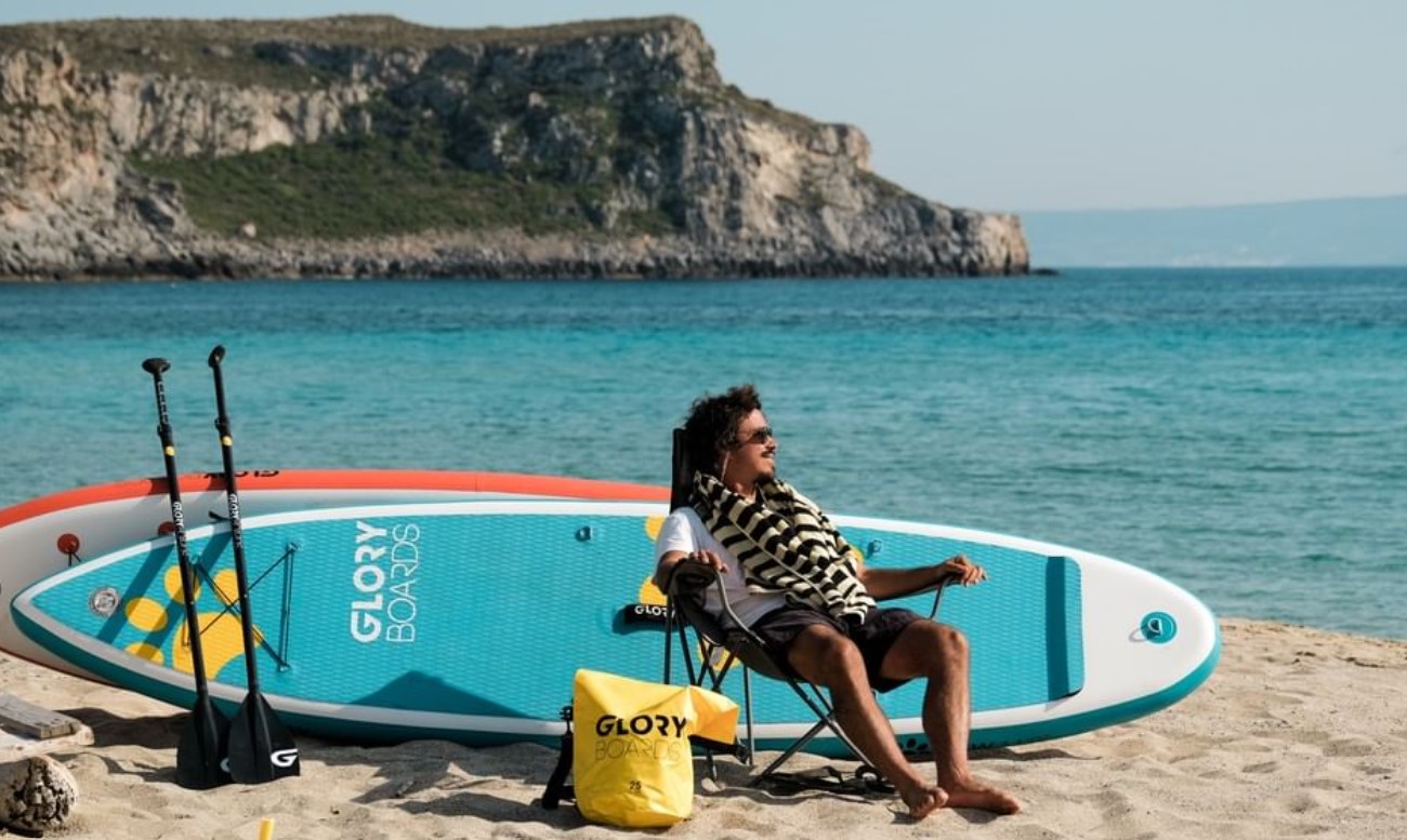 Mann sitz am Strand vor zwei Boards und überlegt ob er Stand Up Paddle Board mieten oder Kaufen soll