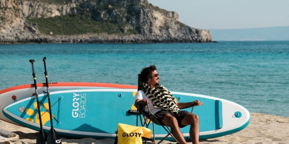 Mann sitz am Strand vor zwei Boards und überlegt ob er Stand Up Paddle Board mieten oder Kaufen soll